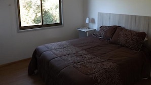 Quarto com uma cama grande, mesa de cabeceira, luminária e janela com vista para árvores.