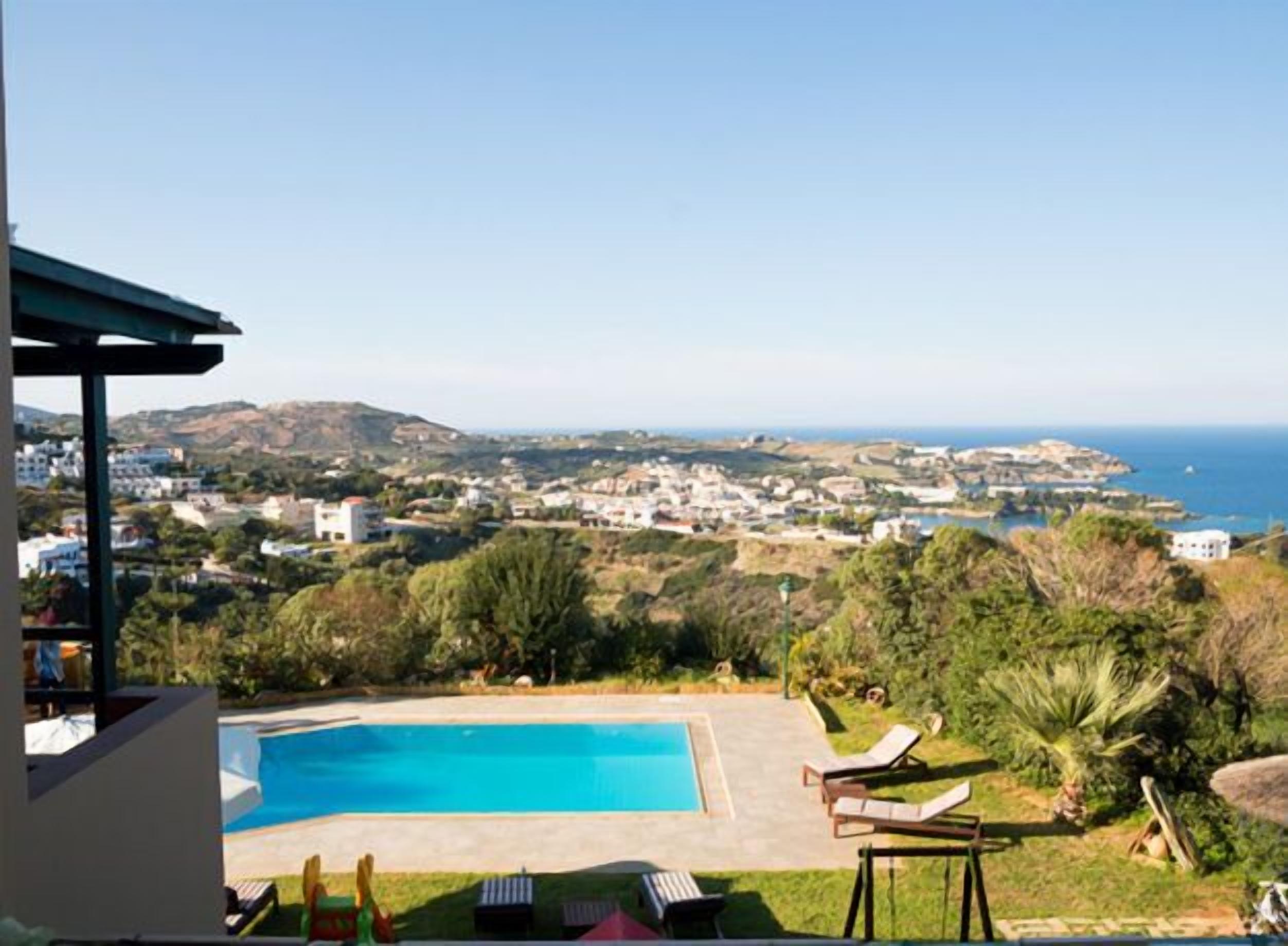 A pool with lounge chairs, a view of a coastal town, and a clear sky.