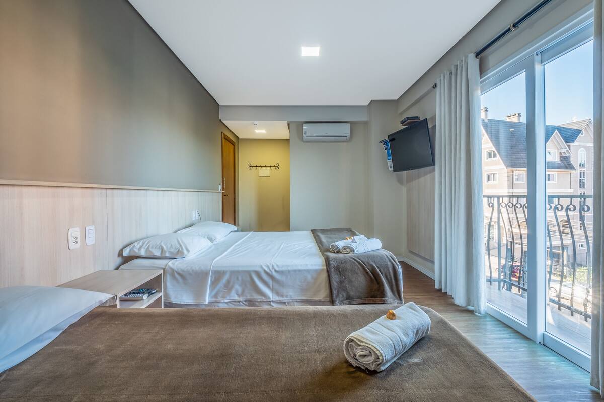 A hotel room with a large bed, a TV mounted on the wall, and a balcony with a view of buildings.