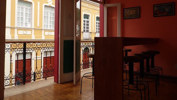 Interior com piso de madeira, parede vermelha e varanda com grades de ferro forjado.