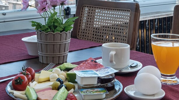 A breakfast table with a variety of food items, a cup of coffee, and a glass of orange juice, set against a backdrop of a city view through large windows. - Oban Suites İstanbul (Istanbul)