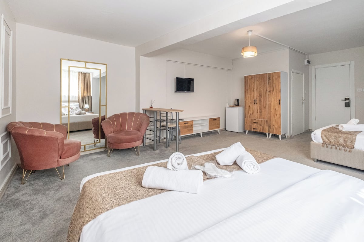 A hotel room with a large bed, two armchairs, a small desk, a TV, a wardrobe, and a mirror.