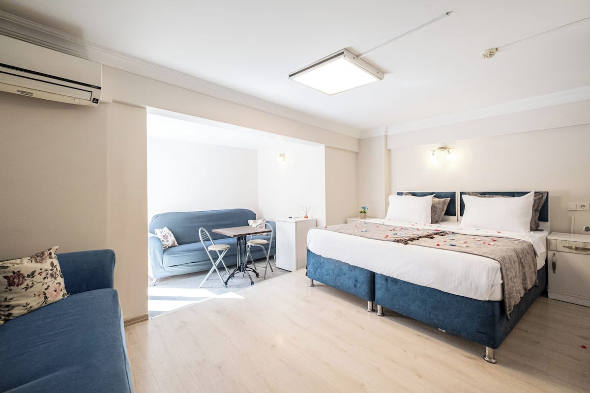 A modern hotel room with a bed, a sofa, a small table, and an air conditioning unit.