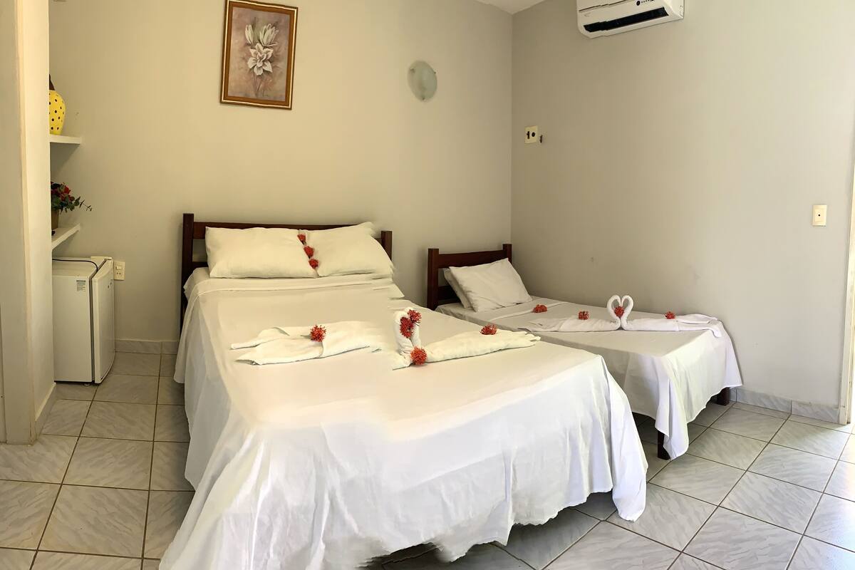 Habitación de hotel con dos camas, cada una con sábanas blancas y flores rojas. Hay un refrigerador pequeño y un equipo de aire acondicionado en la pared.