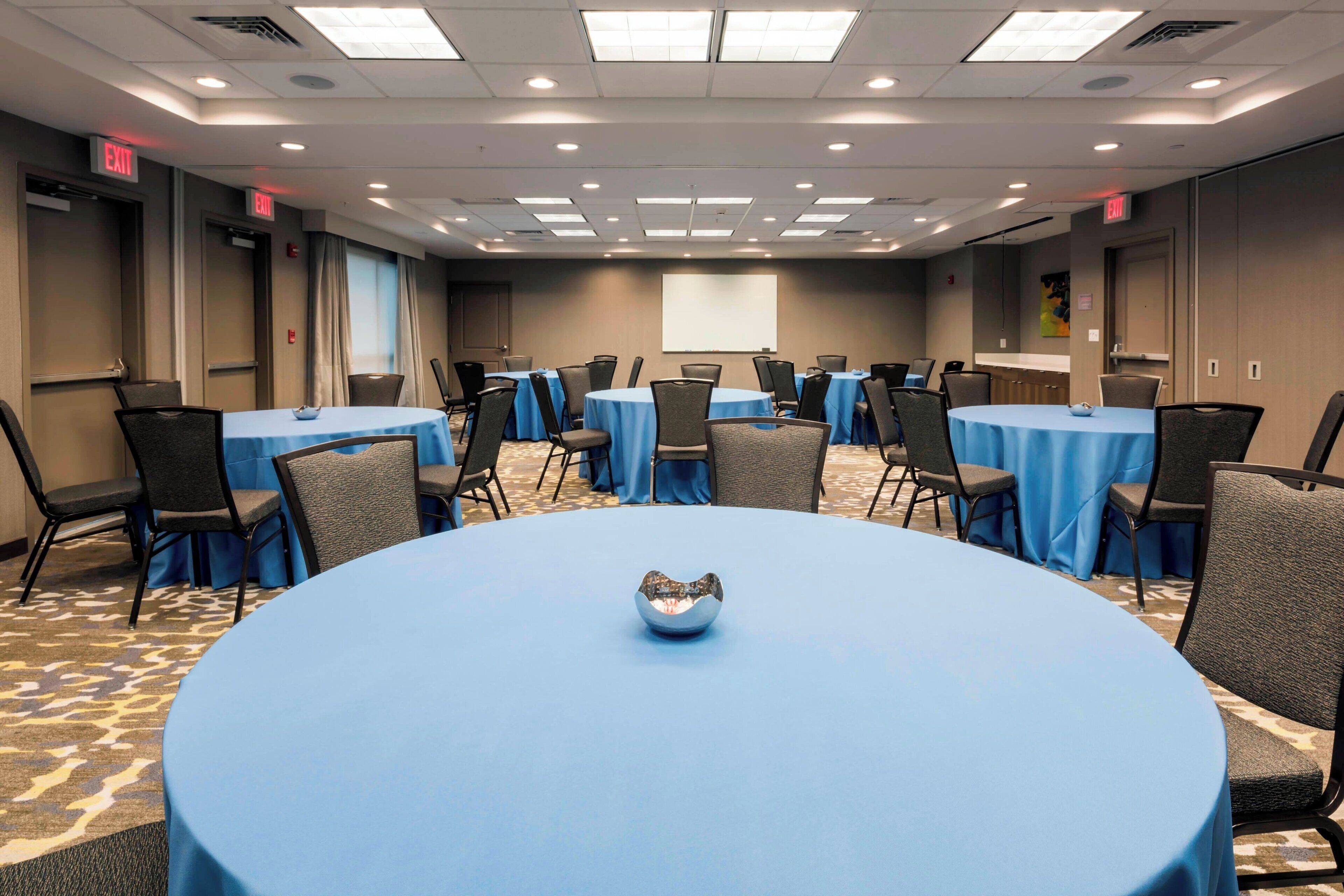 A conference room with multiple tables and chairs, a projector screen, and exit signs.