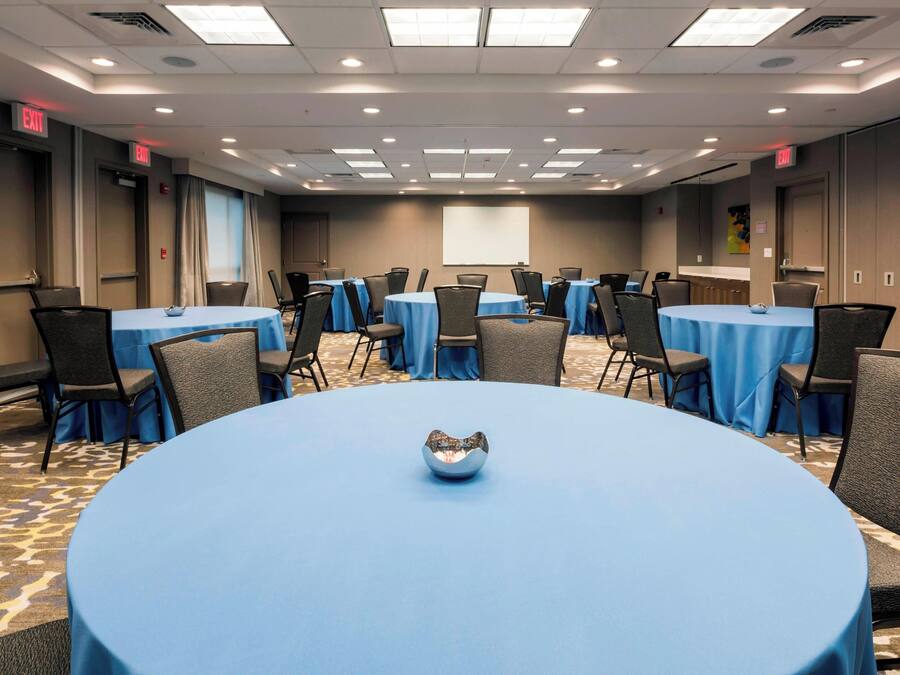 A conference room with multiple tables and chairs, a projector screen, and exit signs.