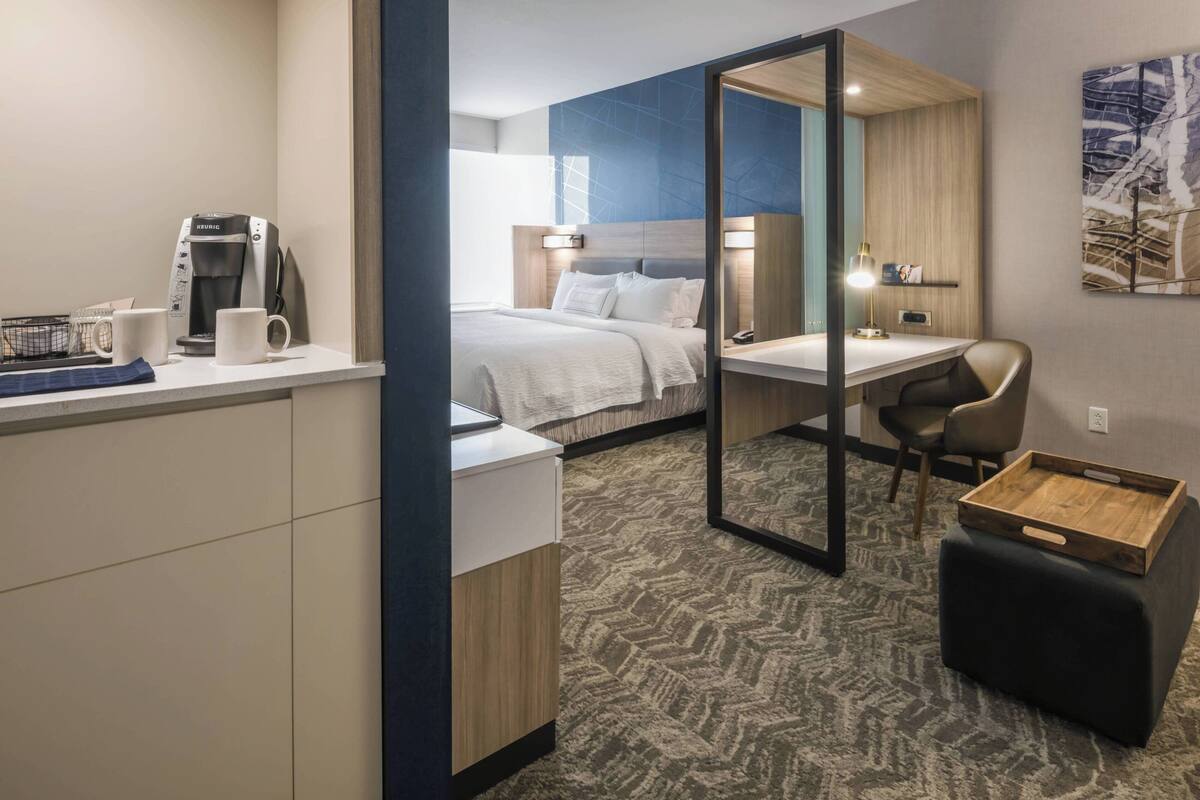 A modern hotel room with a bed, desk, chair, coffee maker, and a small wooden table.