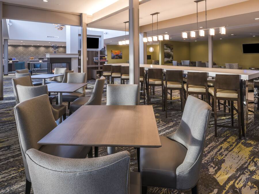 A modern hotel lobby with a central dining area, multiple seating options, and a flat-screen TV mounted on the wall.