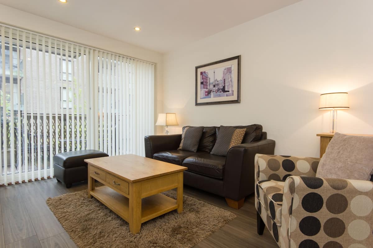 A modern living room with a sofa, a coffee table, and a patterned armchair. There is a window with blinds, a framed picture on the wall, and two lamps on side tables.