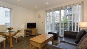 A modern living room with a glass dining table, a wooden coffee table, a leather sofa, a TV on a wooden stand, and large windows with blinds. - Citystay Living - Mill Park Apartments (Cambridge)