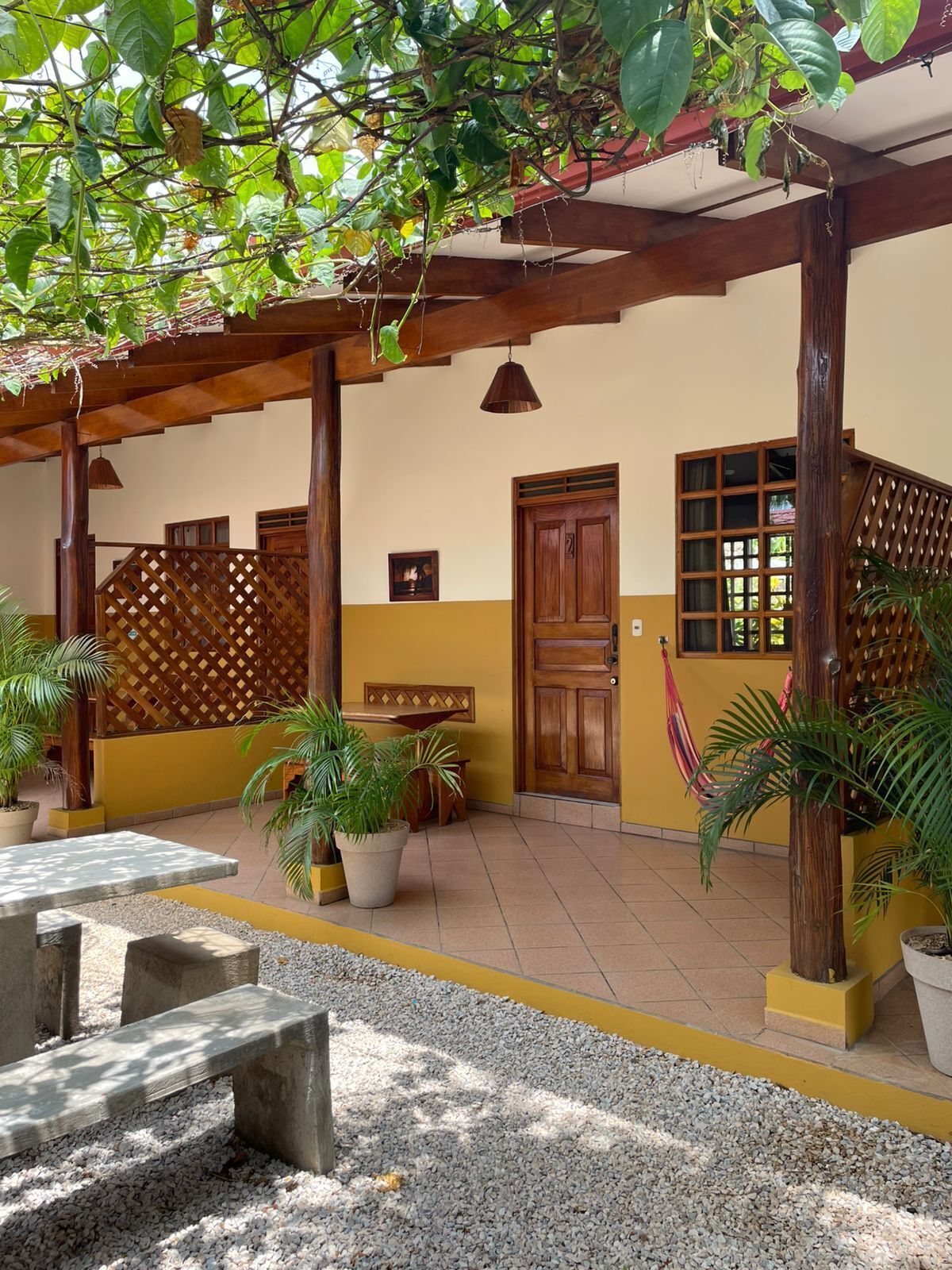 A covered outdoor area with a wooden pergola, potted plants, a bench, and a tiled floor.