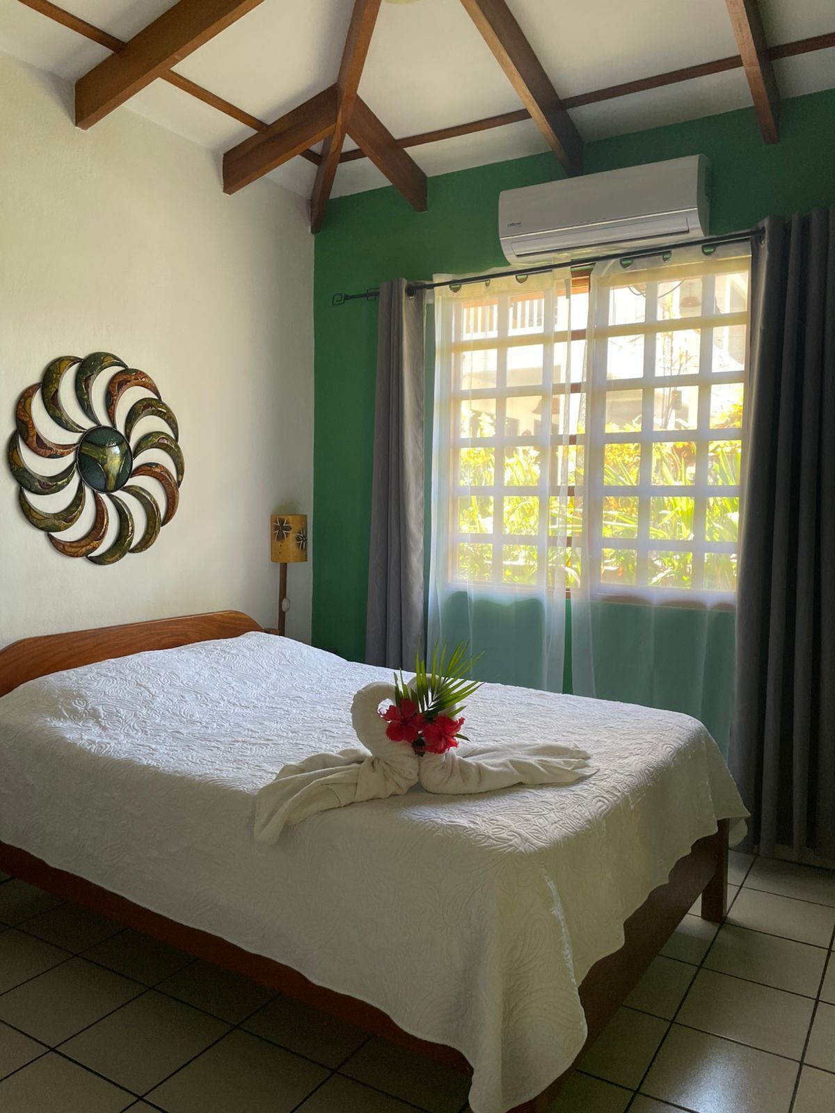 Quarto de hotel com cama, um objeto decorativo na parede, aparelho de ar condicionado e janela com cortinas.