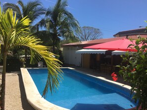 Relax by the serene pool surrounded by lush greenery and palm trees, with a patio area offering shade under a red umbrella.