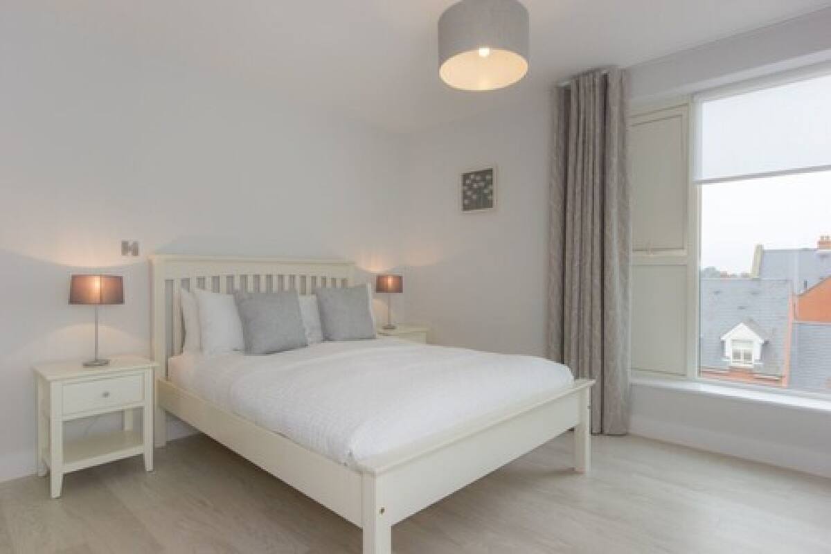 Ein Schlafzimmer mit Bett, Nachttischen, einer Lampe, einem Fenster mit Vorhängen und Blick auf die Stadt.
