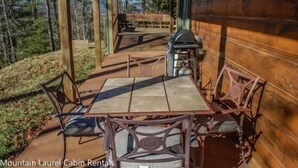 A wooden cabin with a covered outdoor seating area, a barbecue grill, and a stone-tiled table.