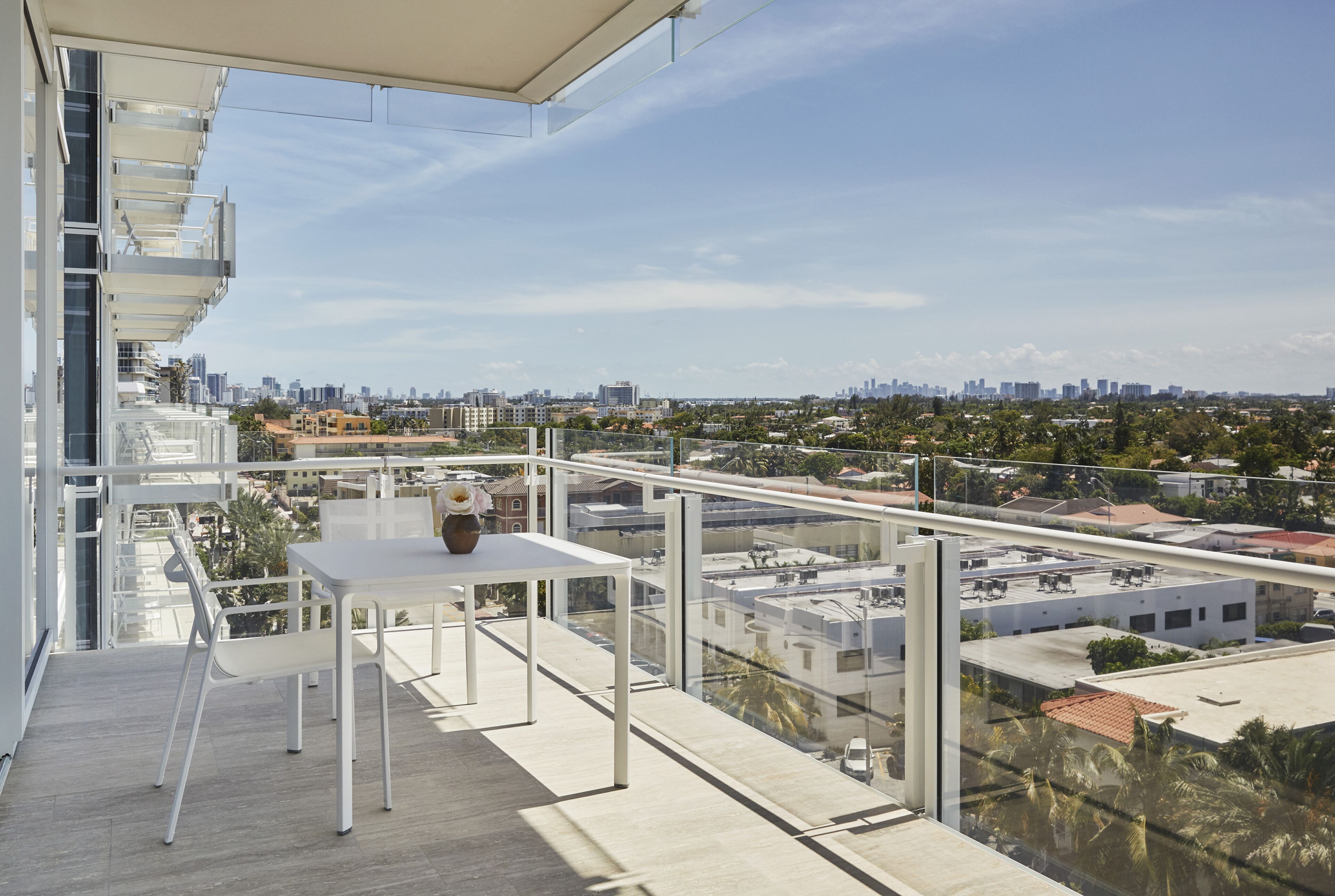 Un balcón con mesa y sillas, con vistas a un paisaje urbano que incluye edificios y zonas verdes.