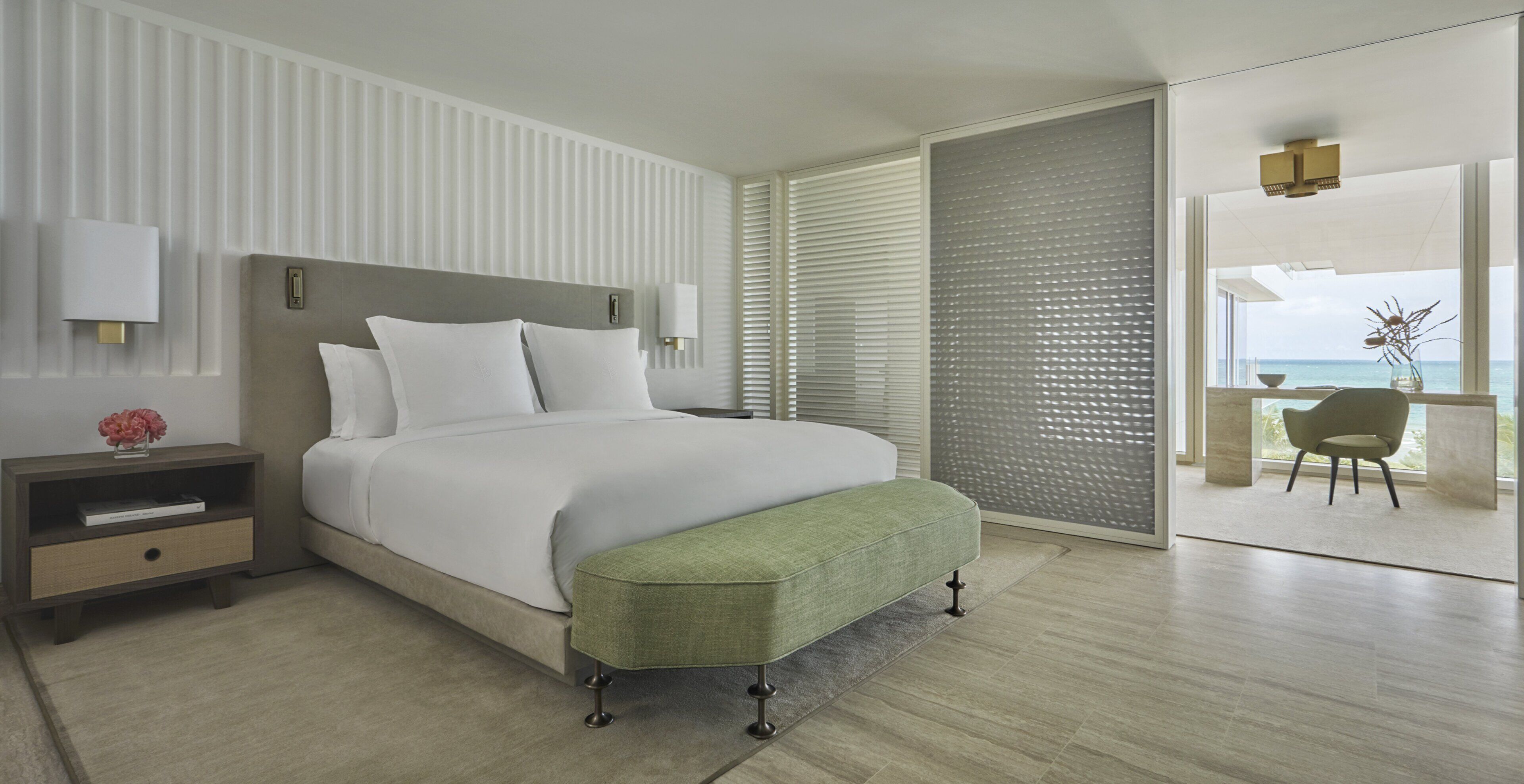 Una habitación de hotel moderna con una cama grande, una mesita de noche, un banco verde, vista al océano y un balcón con una silla.