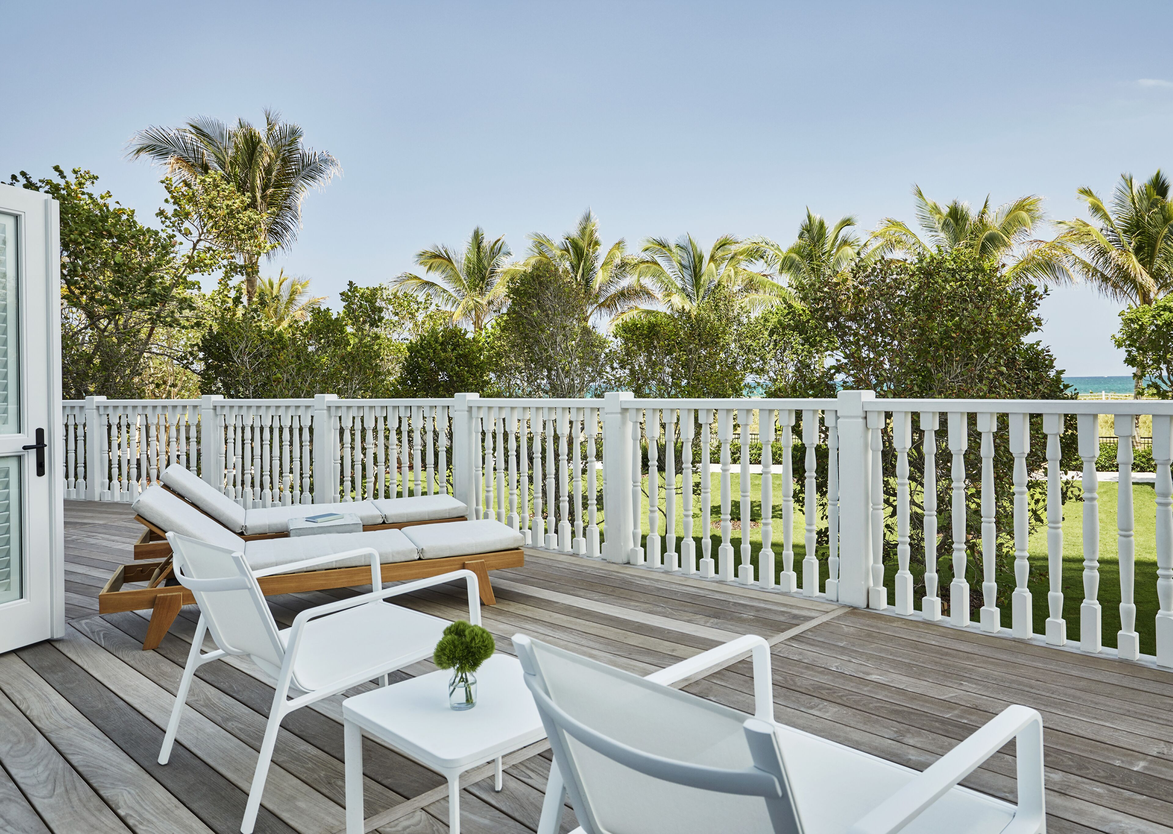Une terrasse dotée de mobilier blanc, comprenant des chaises longues et une table, qui offre une vue sur un jardin luxuriant et des palmiers.
