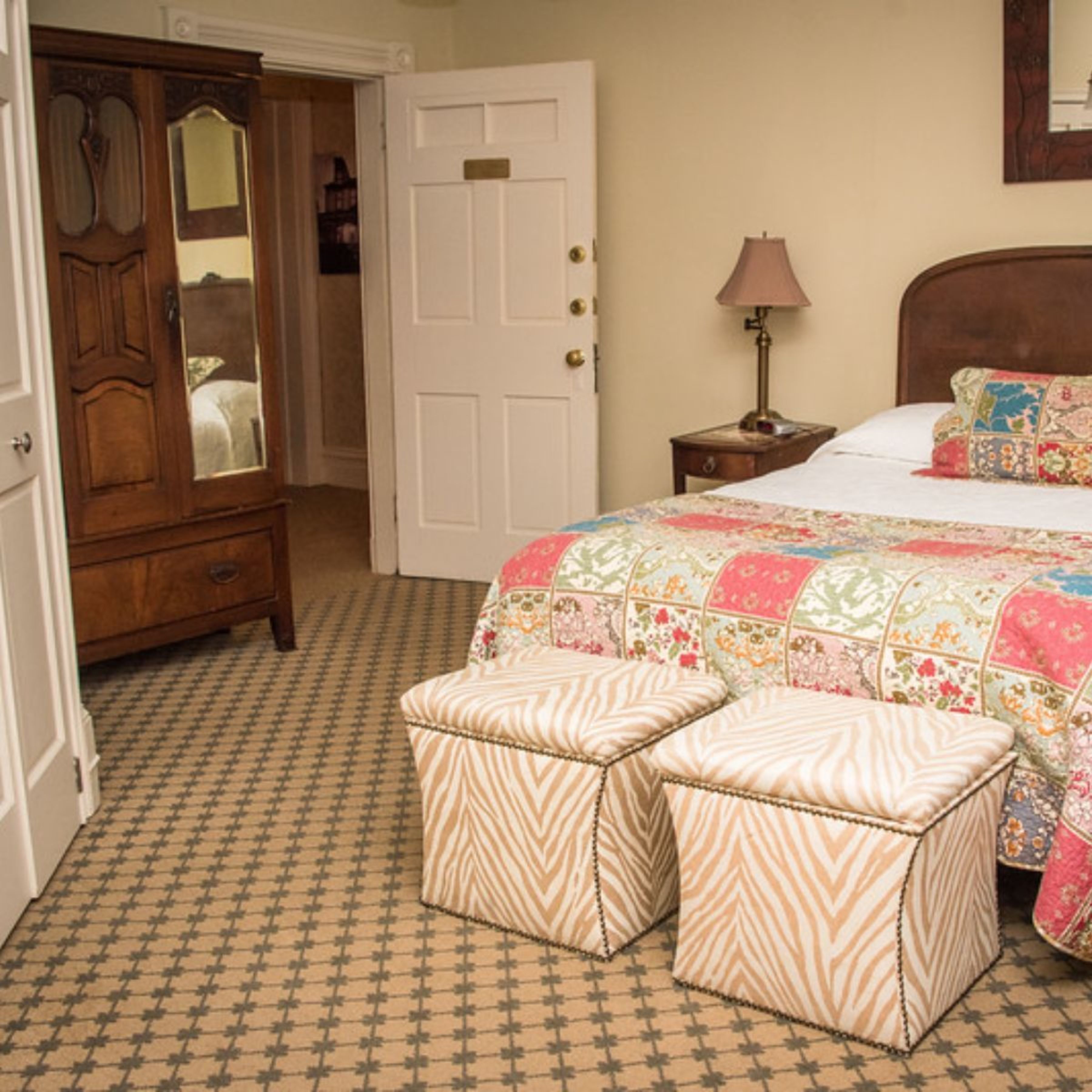 A bedroom with a bed, a dresser, a mirror, a nightstand, and two ottomans.
