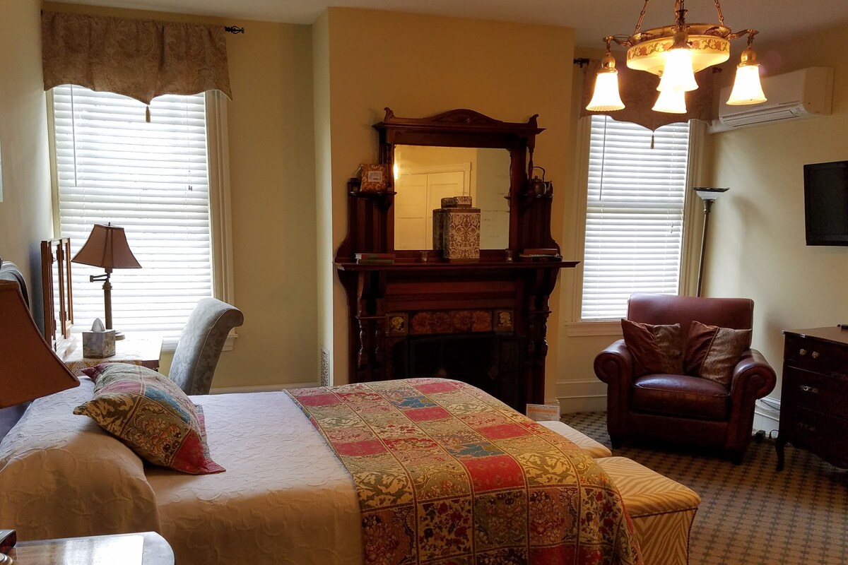A bedroom with a bed, a chair, a dresser, a mirror, and a fireplace.