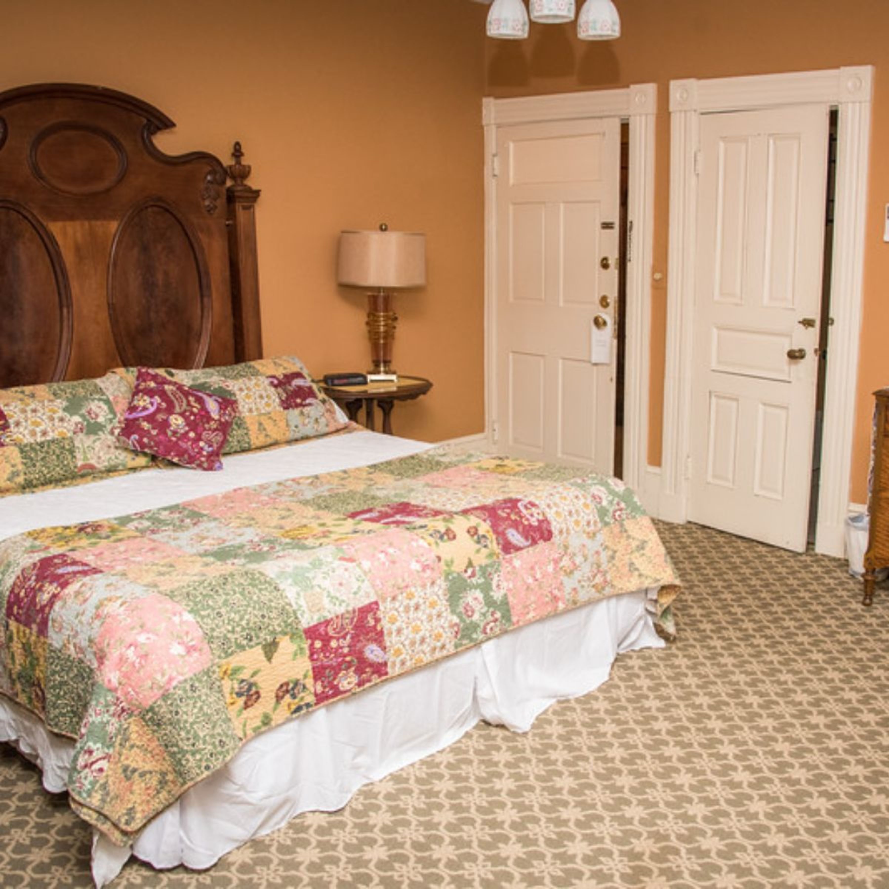 A bedroom with a large bed, a wooden headboard, a bedside table with a lamp, and two white doors.