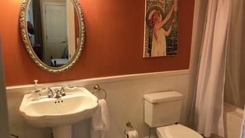 Une salle de bain avec un lavabo blanc, des toilettes et un grand miroir. Il y a un tableau au mur et un rideau de douche.