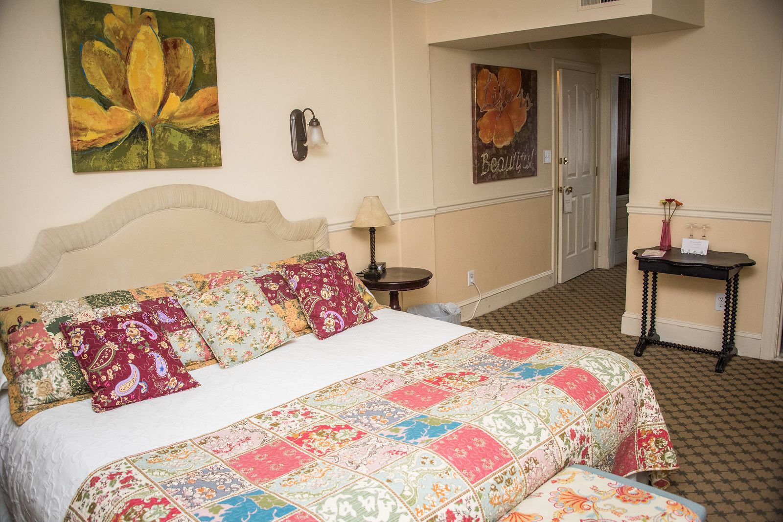 A neatly made bed with a floral quilt, a wall-mounted lamp, and a painting of flowers.