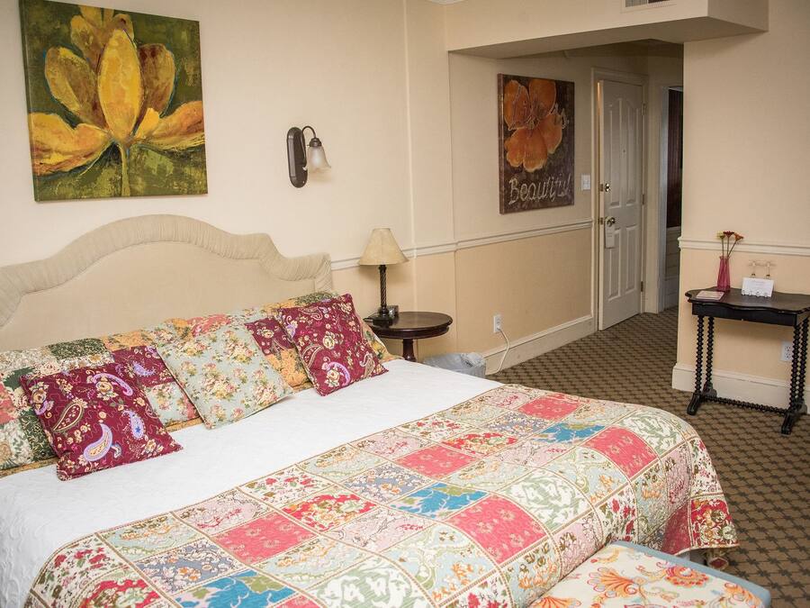 Un lit bien fait, recouvert d’une courtepointe à motifs floraux, une lampe fixée au mur et un tableau représentant des fleurs.