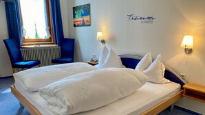 Una cama doble con sábanas y almohadones blancos, una silla azul, una mesita de noche con una lámpara, y una pared con un cuadro de un paisaje urbano y un cartel con un nombre.