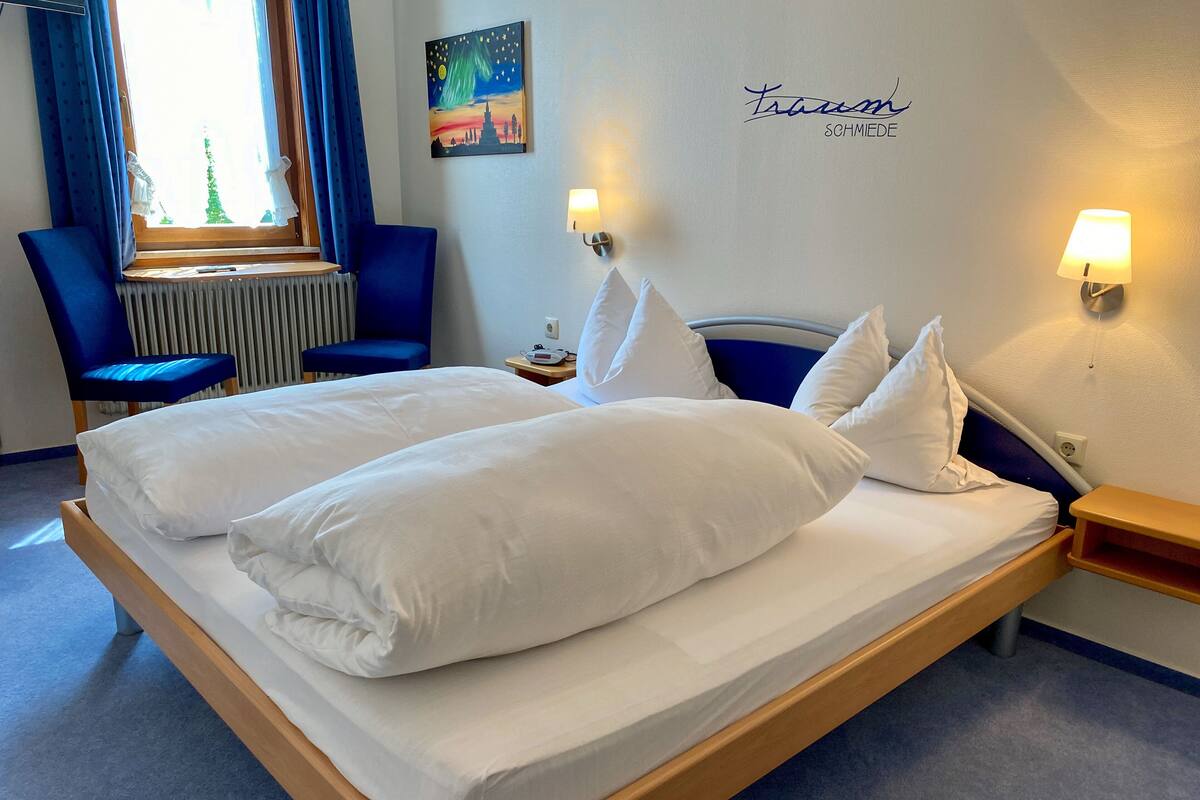 A double bed with white linens and pillows, a blue chair, a nightstand with a lamp, and a wall with a cityscape painting and a name sign.
