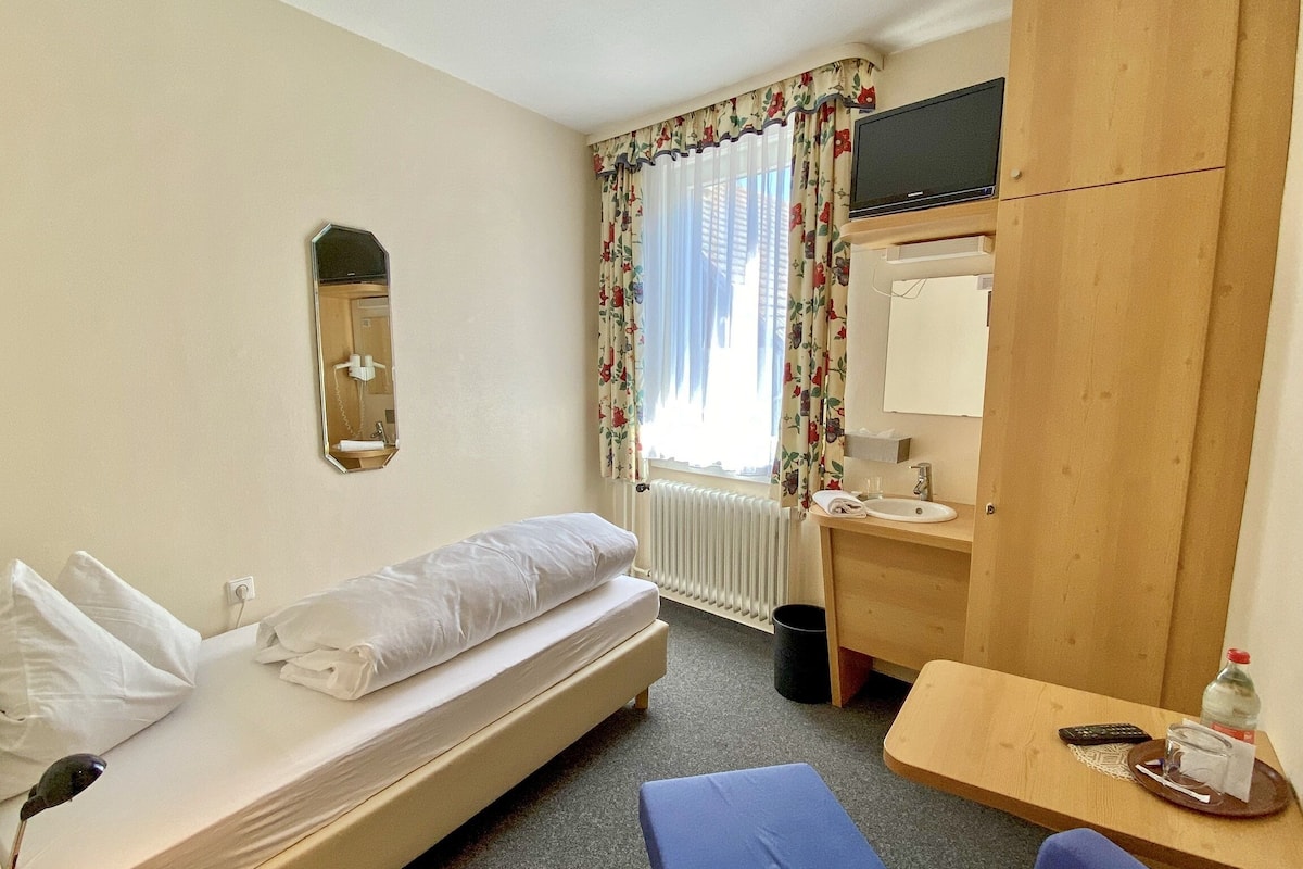 A hotel room with a bed, a small desk with a chair, a mirror, a television, and a window with curtains.