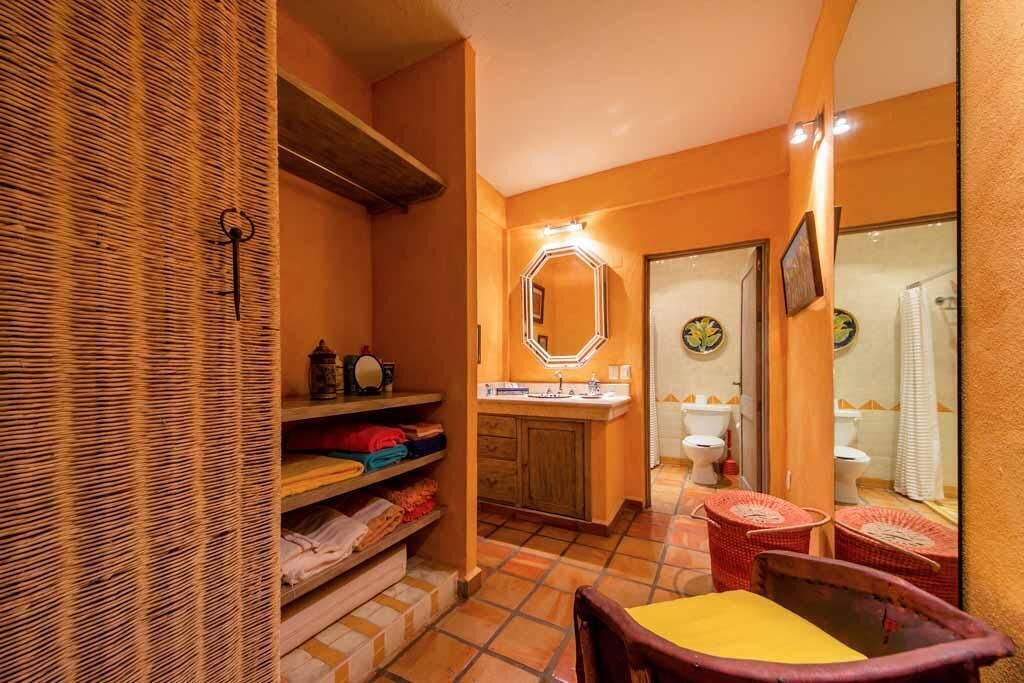 A compact bathroom with a tiled floor, a wooden shelf with folded towels, a mirror with lights, and a small seating area with a yellow cushion.