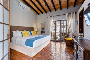 Un dormitorio con una cama grande, techo de madera, suelo de baldosas, un balcón con una silla y un aparador.