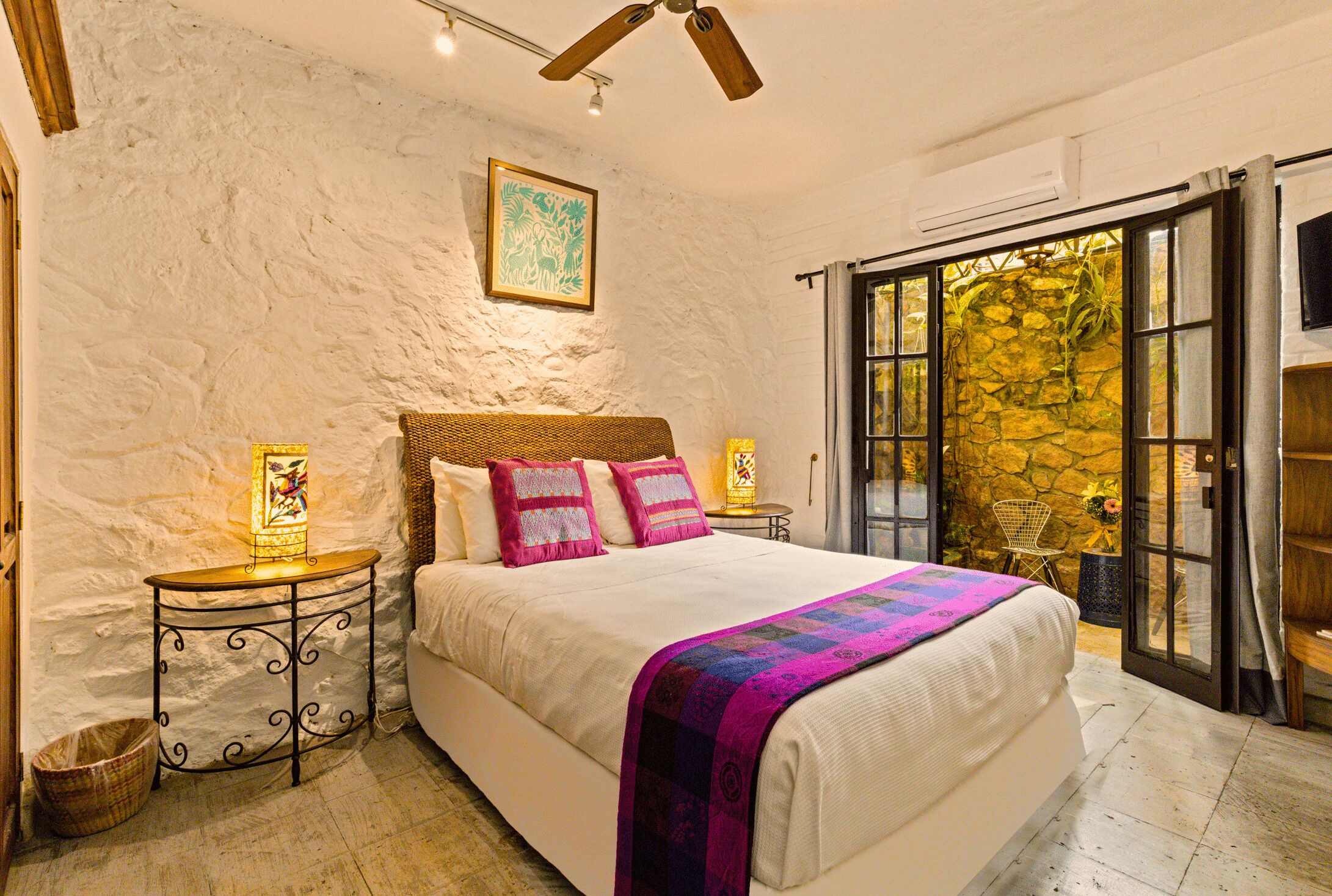A bedroom with a large bed, a stone wall, a wooden ceiling fan, and a view through a glass door.