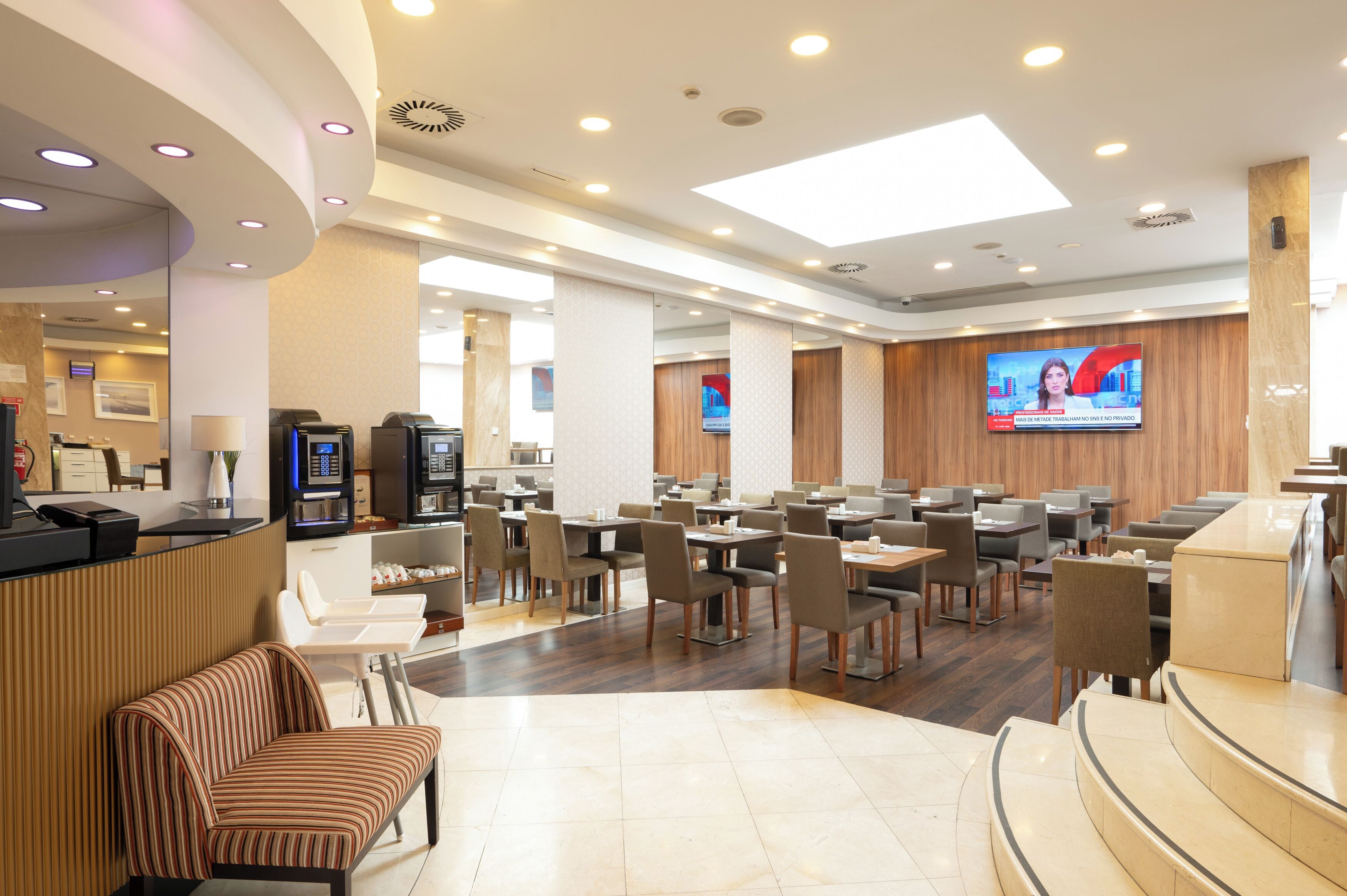 Una hall d'albergo moderna con reception, area salotto e uno schermo televisivo che mostra un notiziario.