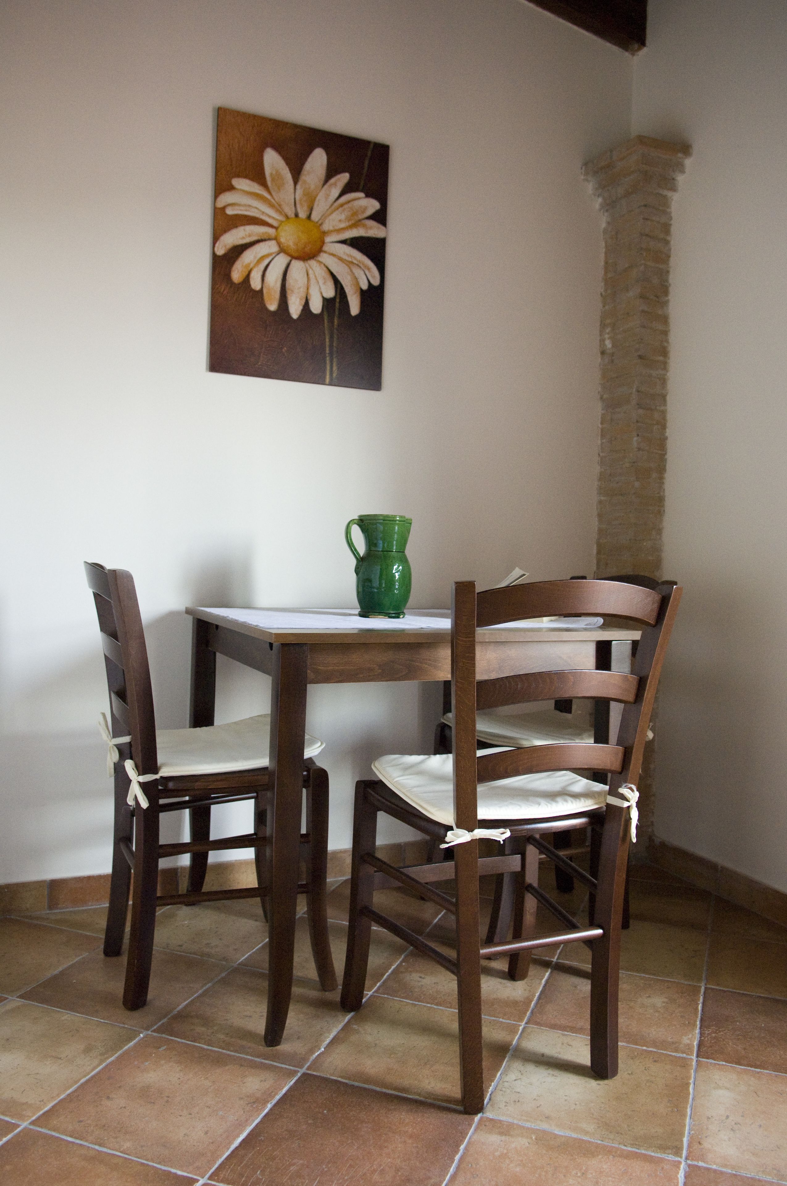 A dining area with a wooden table and chairs, a green pitcher, and a painting of a daisy on the wall.
