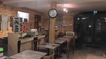 A cozy restaurant interior with wooden walls, multiple tables and chairs, a counter with various items, and a clock on the wall.