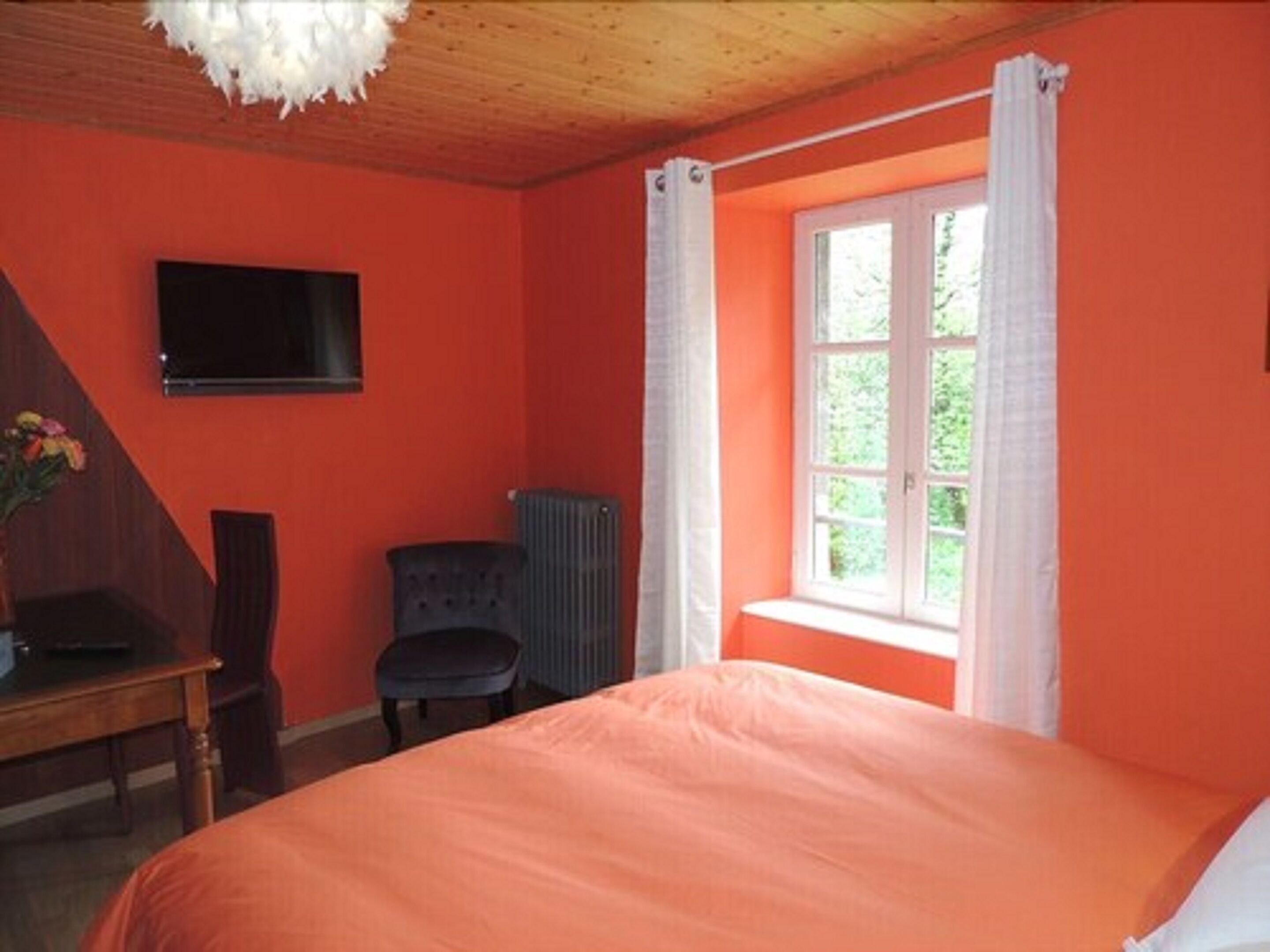 Un dormitorio con una cama, televisión montada en la pared, radiador y ventana con cortinas.