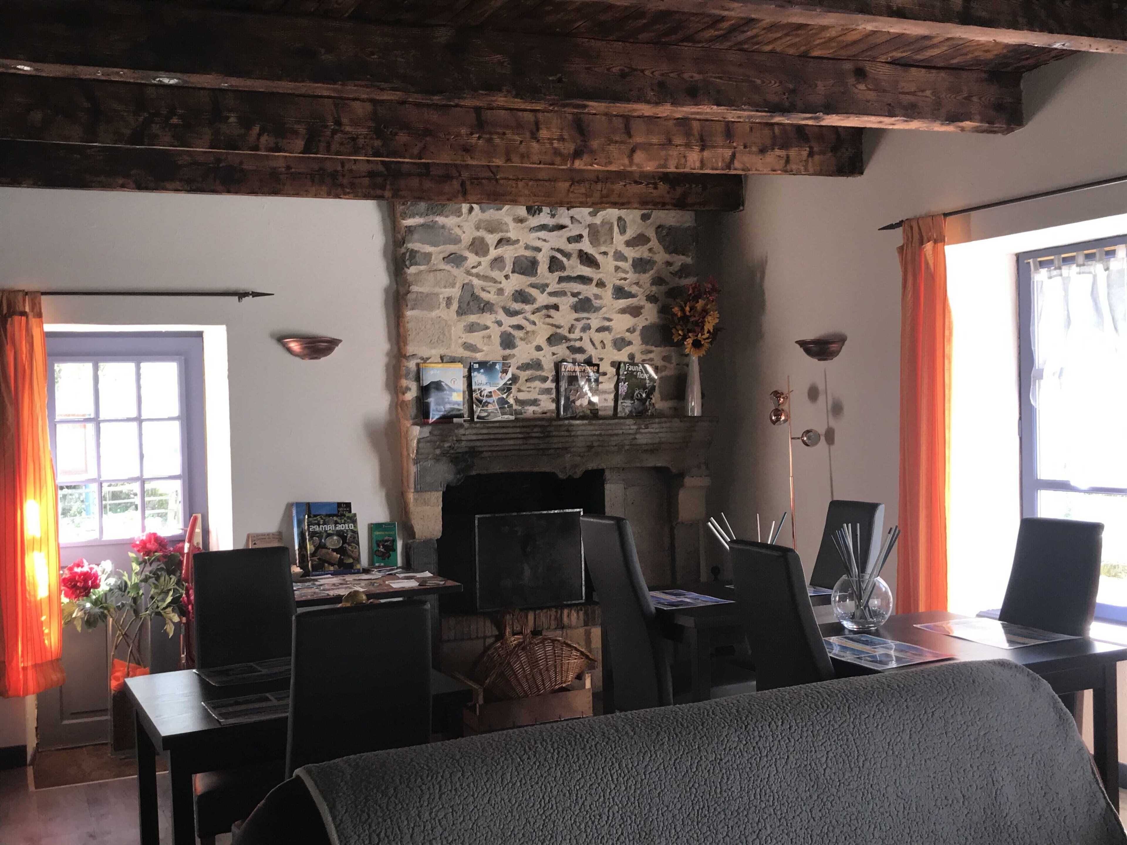 A rustic room with a stone fireplace, wooden beams, a dining table with chairs, and a window with curtains.