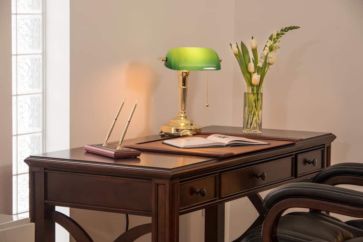 A wooden desk with a green desk lamp, a book, a pen, and a vase with flowers.
