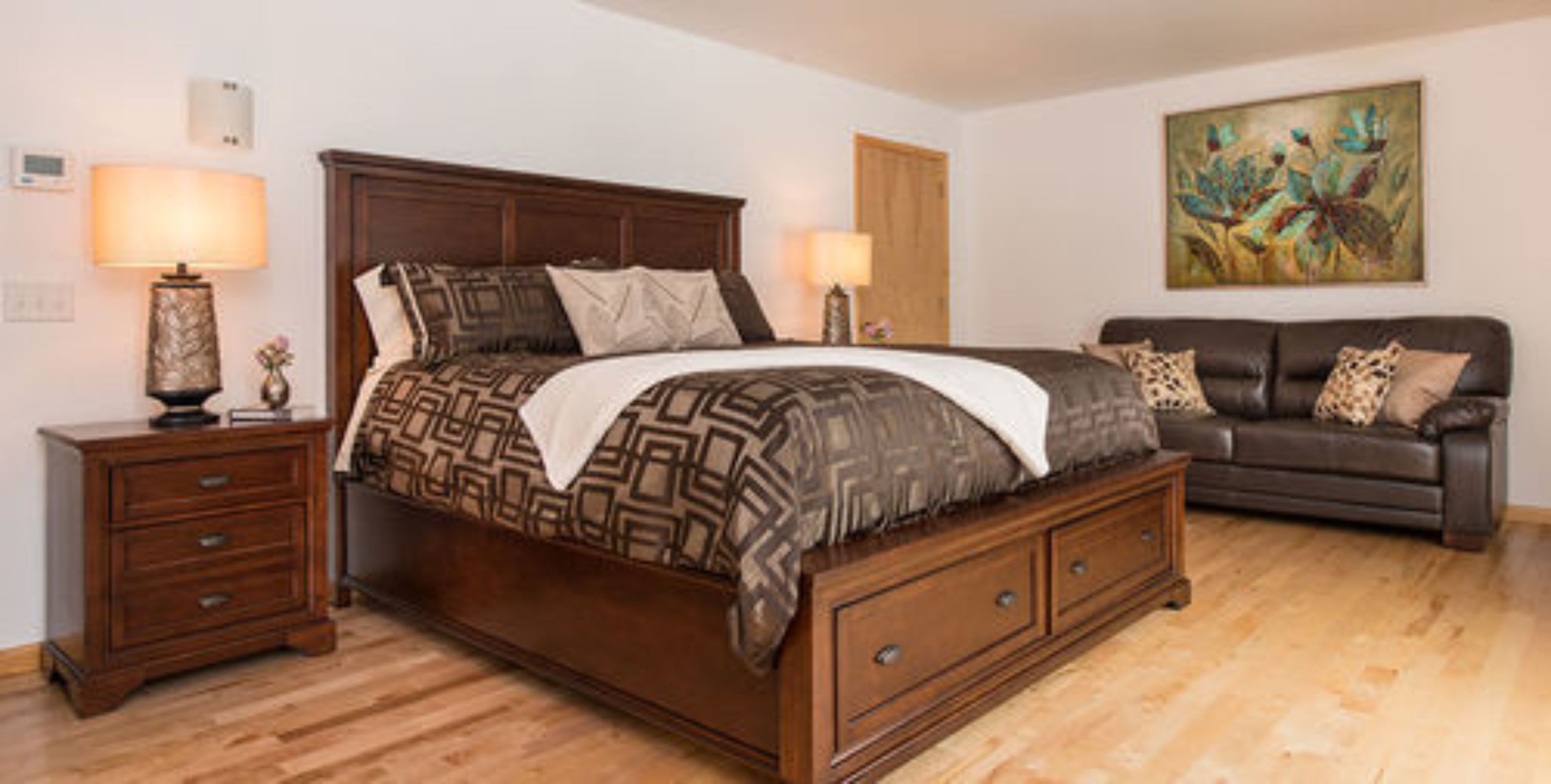 A bedroom with a large bed, two bedside tables, a dresser, a dark brown leather sofa, and a painting on the wall.