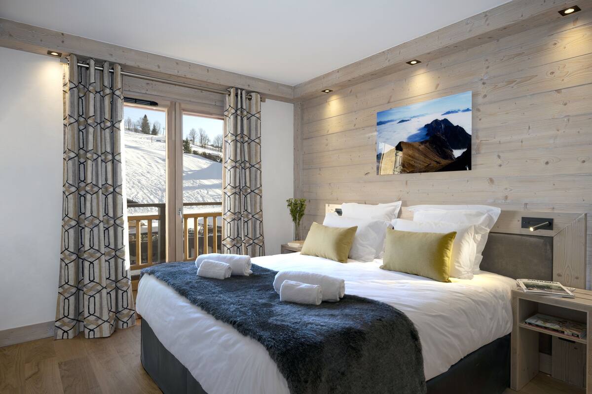 A modern bedroom with a large bed, wooden walls, a view of a snowy landscape, and a painting of mountains.