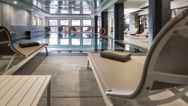 Een modern binnenzwembad met lounge fauteuils en een glazen wand.