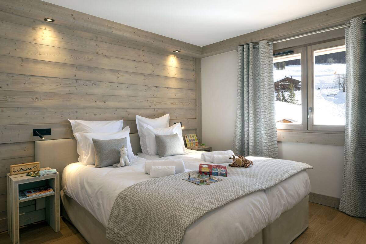 A bedroom with a wooden headboard, a bed with white bedding, a nightstand with a lamp, and a window with a view of a snowy landscape.