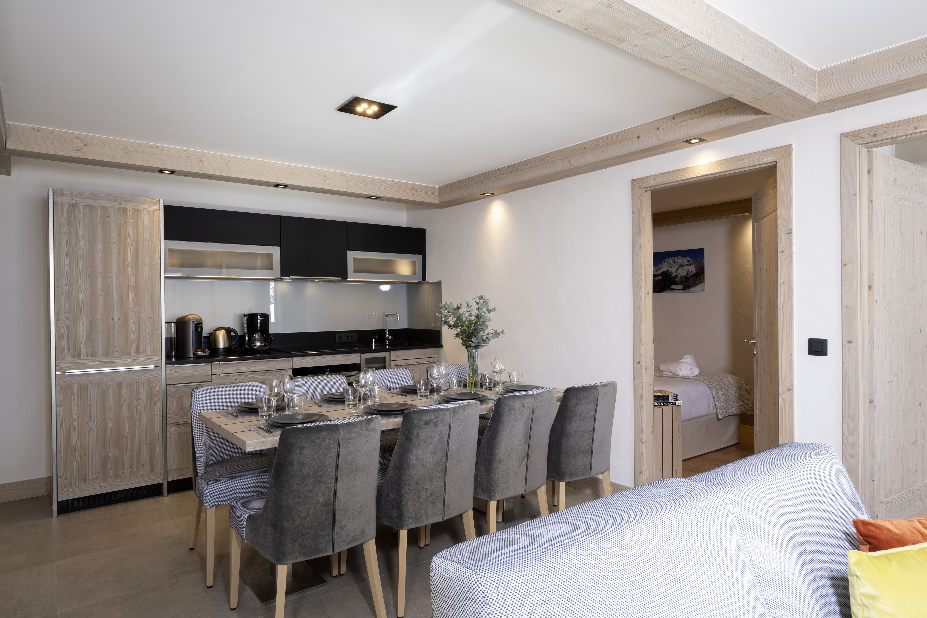 A modern kitchen with a dining area, wooden beams on the ceiling, and a bed in the adjacent room.