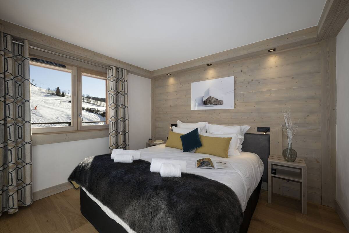 A bedroom with a large bed, wooden walls, a window with a view of a snowy landscape, and a small bedside table with a vase.