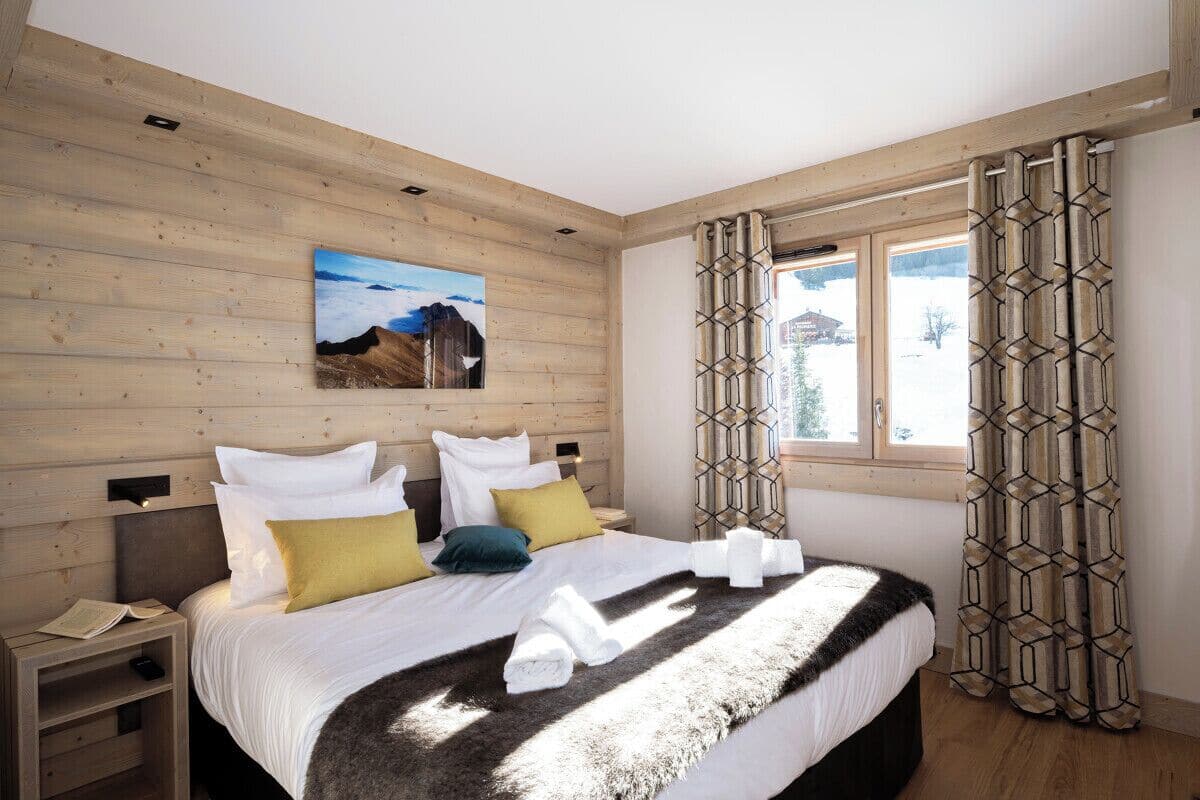 A bedroom with a wooden wall, a bed with white bedding, a nightstand, and a window with patterned curtains.