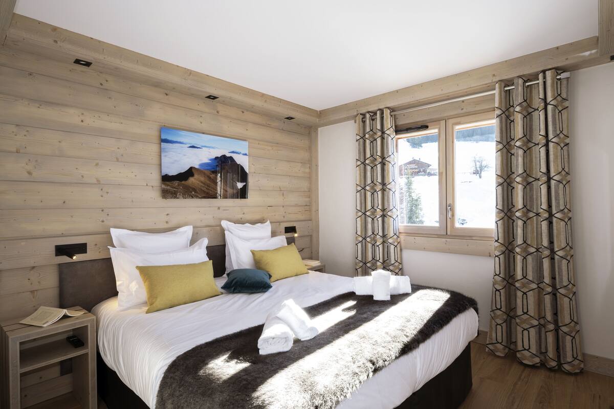 A hotel room with a wooden ceiling, a bed with white bedding and multiple pillows, a nightstand with a book and remote, and a window with patterned curtains.