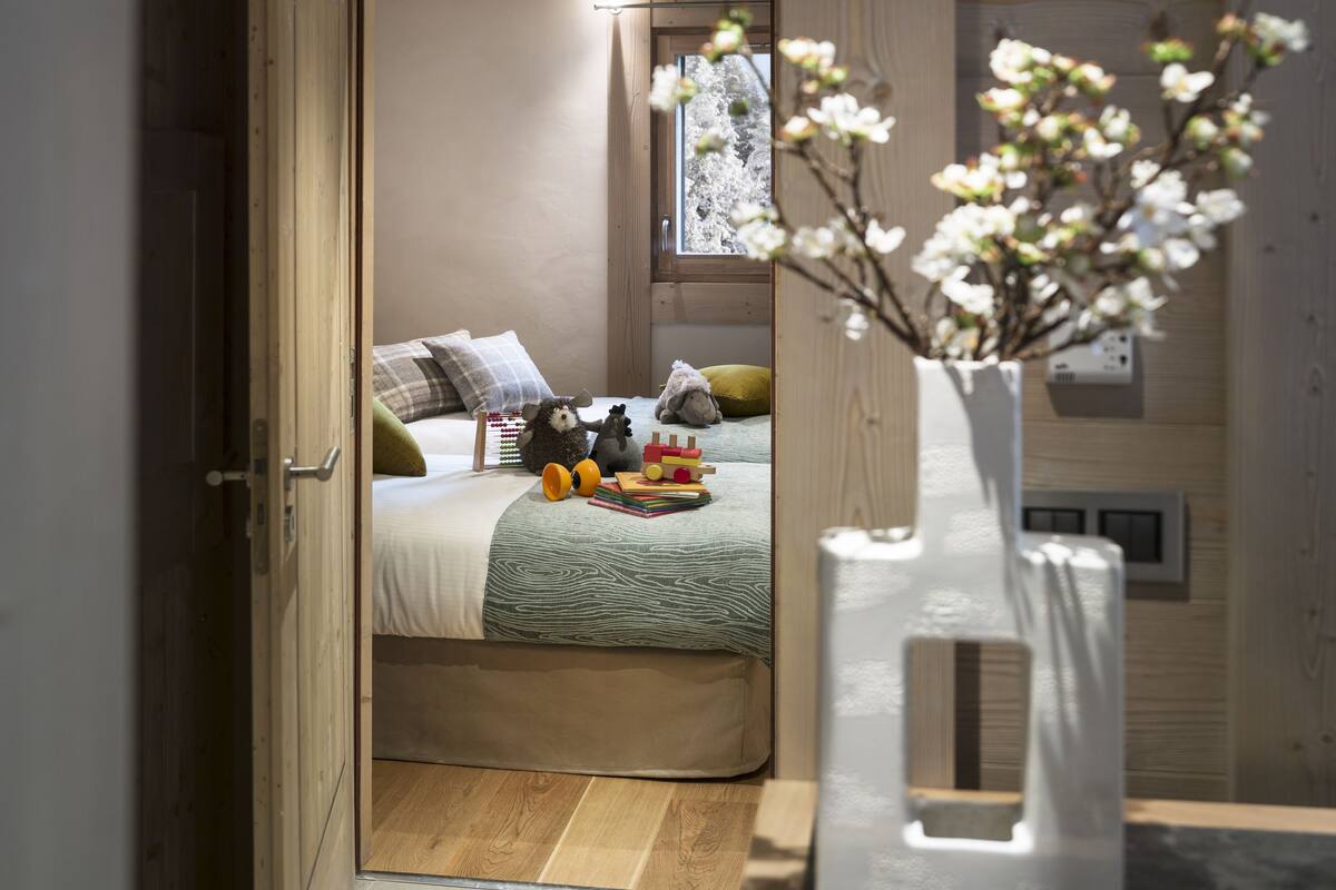 A bedroom with a bed, pillows, a nightstand, a vase with flowers, and a window with a view of trees.