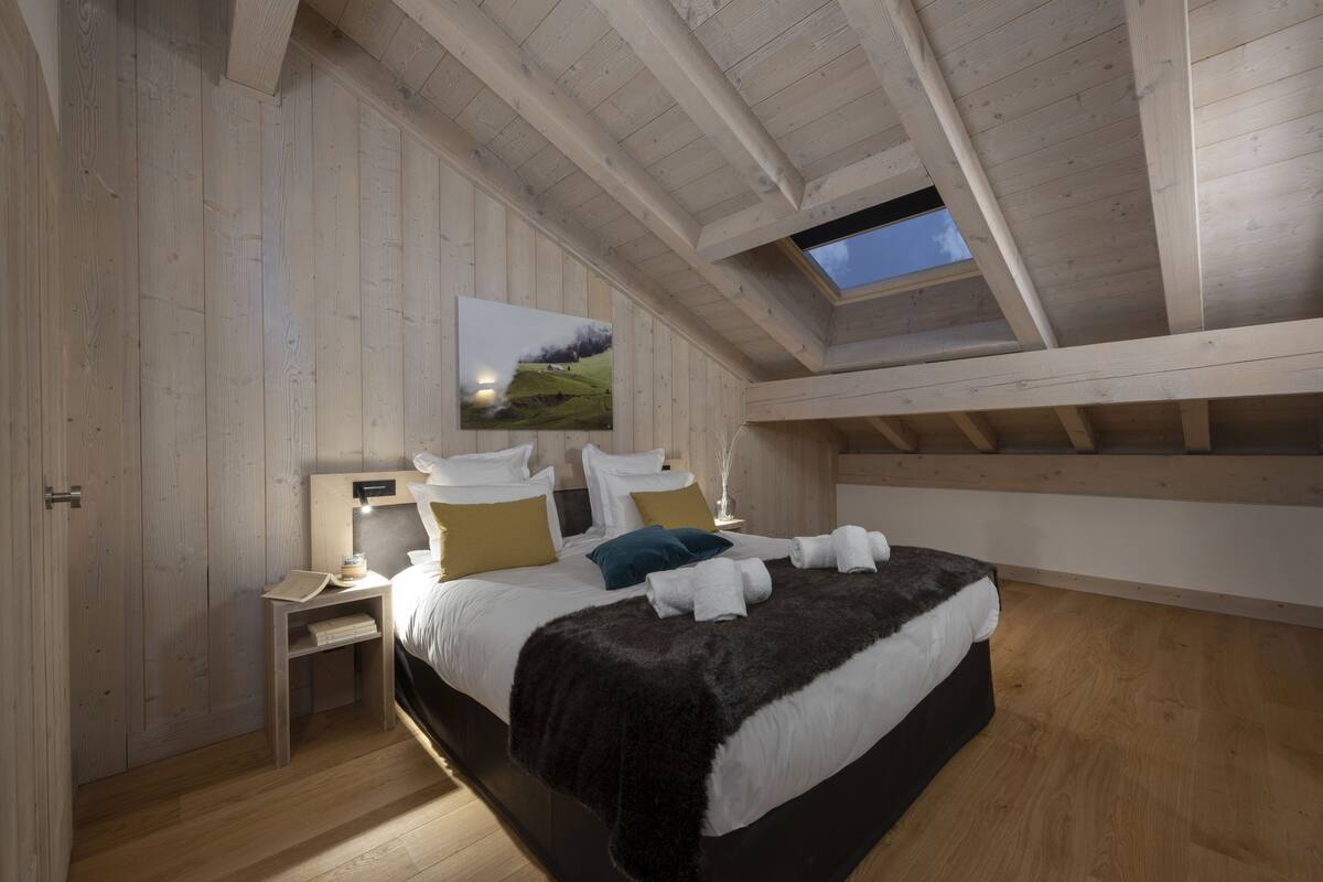 A bedroom with a wooden ceiling, a skylight, a bed with white bedding and a dark throw, a nightstand with a lamp, and a painting on the wall.
